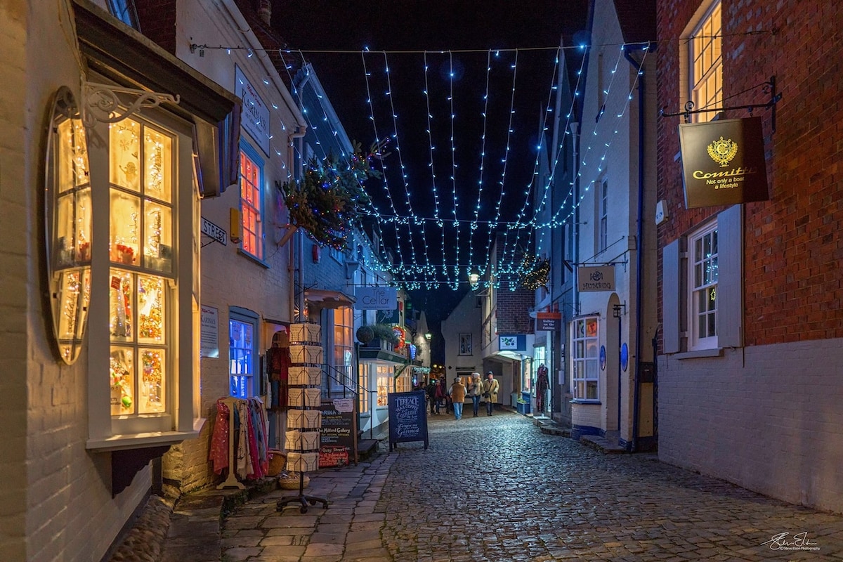 A historic cobbled street is illuminated by twinkling string lights overhead, creating a charming ambiance. Shops with inviting window displays line both sides, blending vintage and modern aesthetics. Visitors can be seen strolling along the path, enjoying the vibrant atmosphere of the area.
