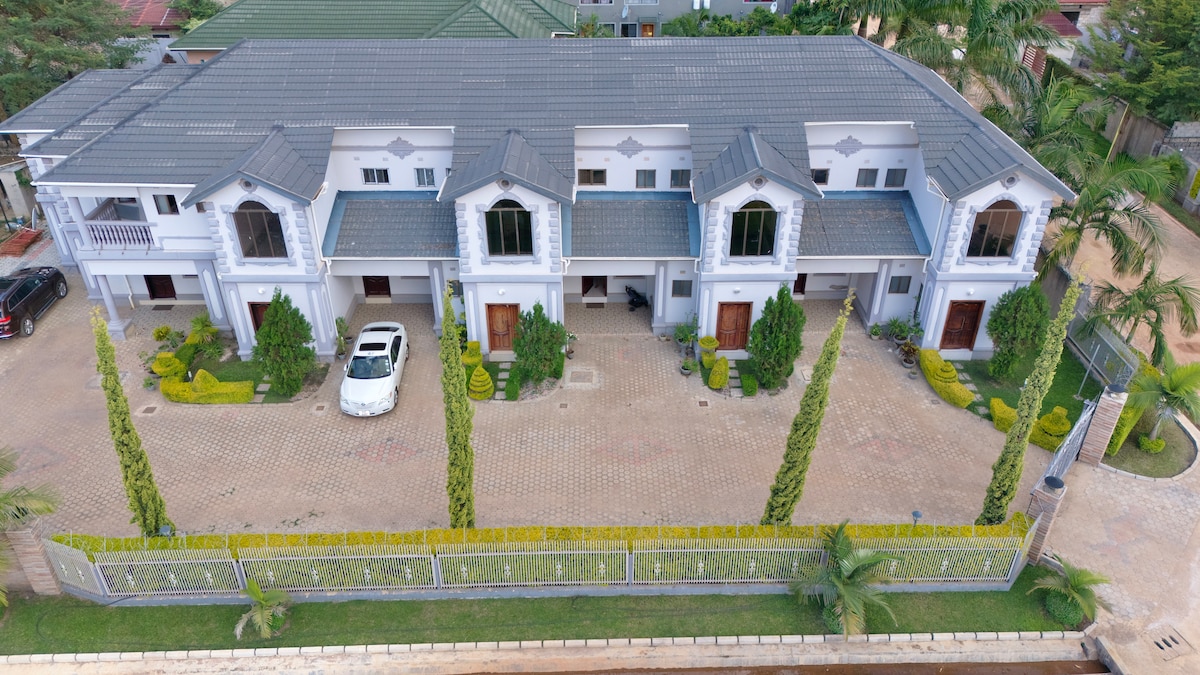 An aerial view of the Lapaz Apartments displays a modern two-story building with a symmetrical façade. Each unit features distinct entrances framed by shrubbery and decorative plants. The driveway accommodates parked vehicles, and the surrounding landscape includes neatly trimmed hedges and palm trees.
