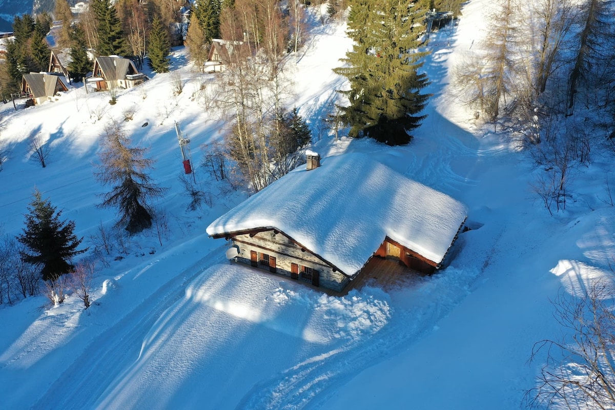 An aerial view reveals a spacious chalet covered in fresh snow, surrounded by trees. The stone exterior blends harmoniously with the winter landscape, and a sloping snowy path leads to the entrance, enhancing the secluded ambiance of this peaceful mountain retreat.