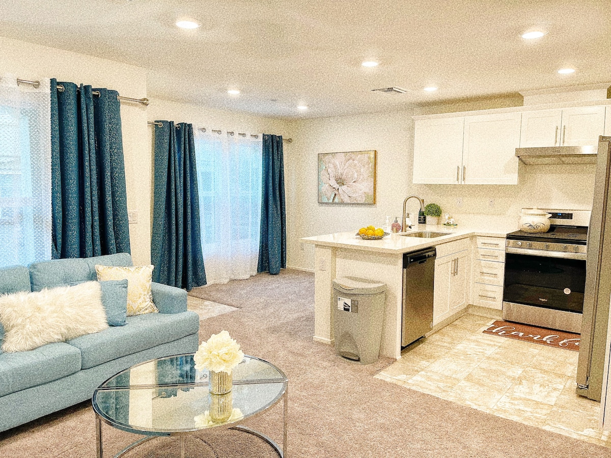 A modern kitchen and living area are showcased, featuring a light blue sofa with decorative pillows and a glass coffee table. The kitchen includes white cabinetry and stainless-steel appliances. Natural light filters through sheer curtains, illuminating the plush carpeted floor.