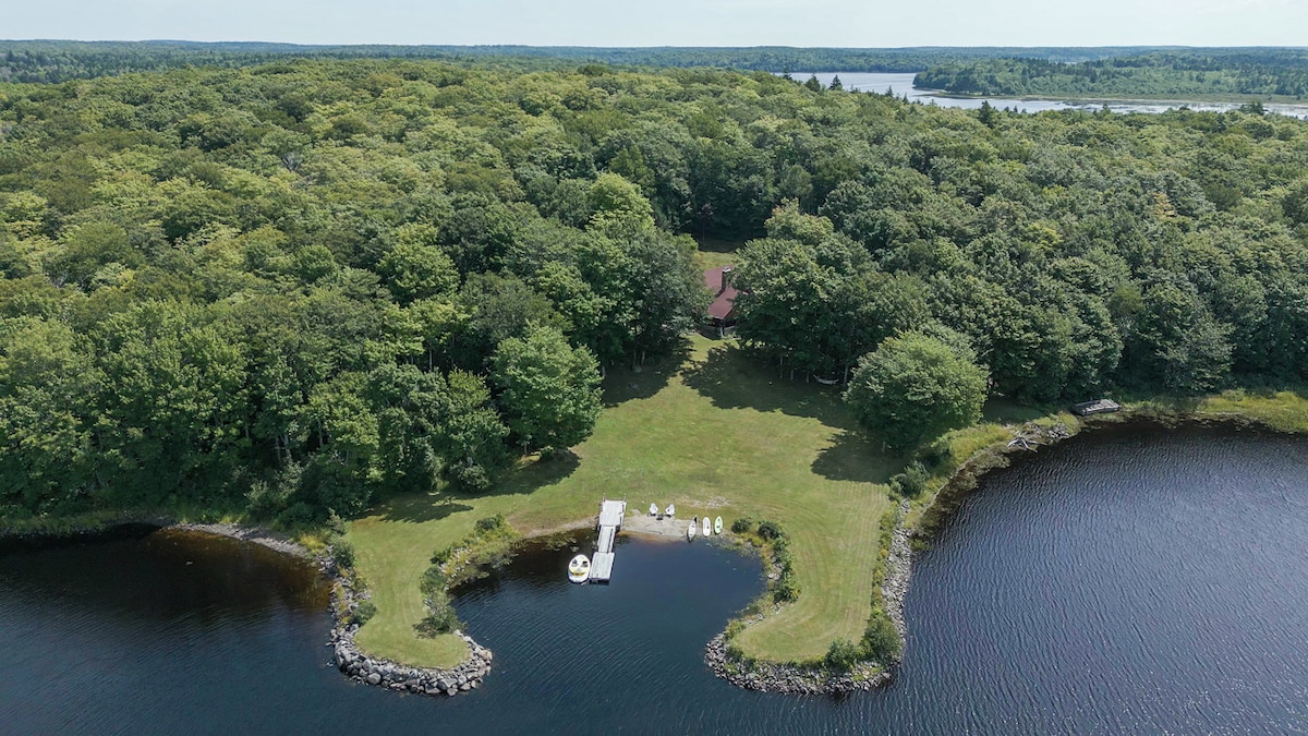 An aerial view showcases the expansive green lawn of the property bordered by the lake. Tall trees provide a natural backdrop, while a dock extends into the water. The surrounding landscape features a serene waterway and dense forest, conveying a sense of seclusion.
