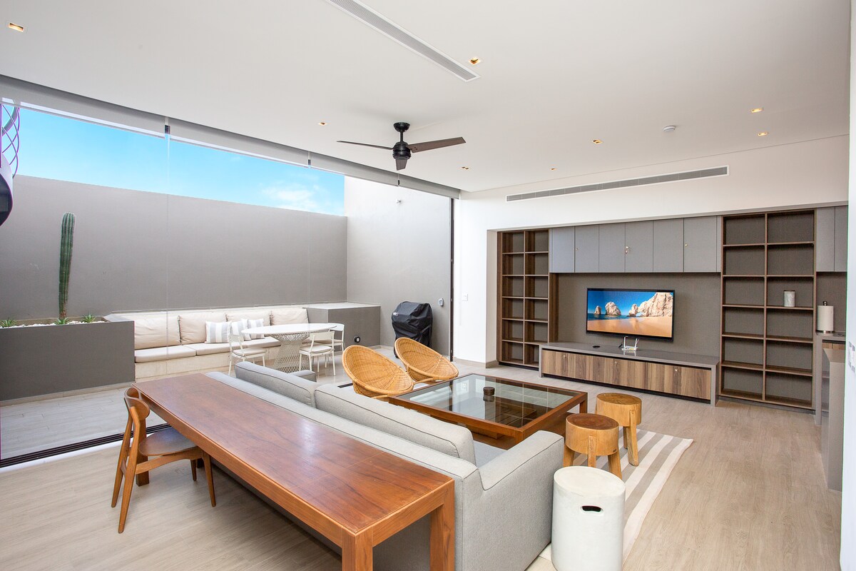 A spacious living area features large glass doors that open to an outdoor seating area. Comfortable furniture includes a sectional sofa and a wooden dining table. A flat-screen TV is mounted on a neutral-toned wall, while natural light brightens the space, enhancing its openness.