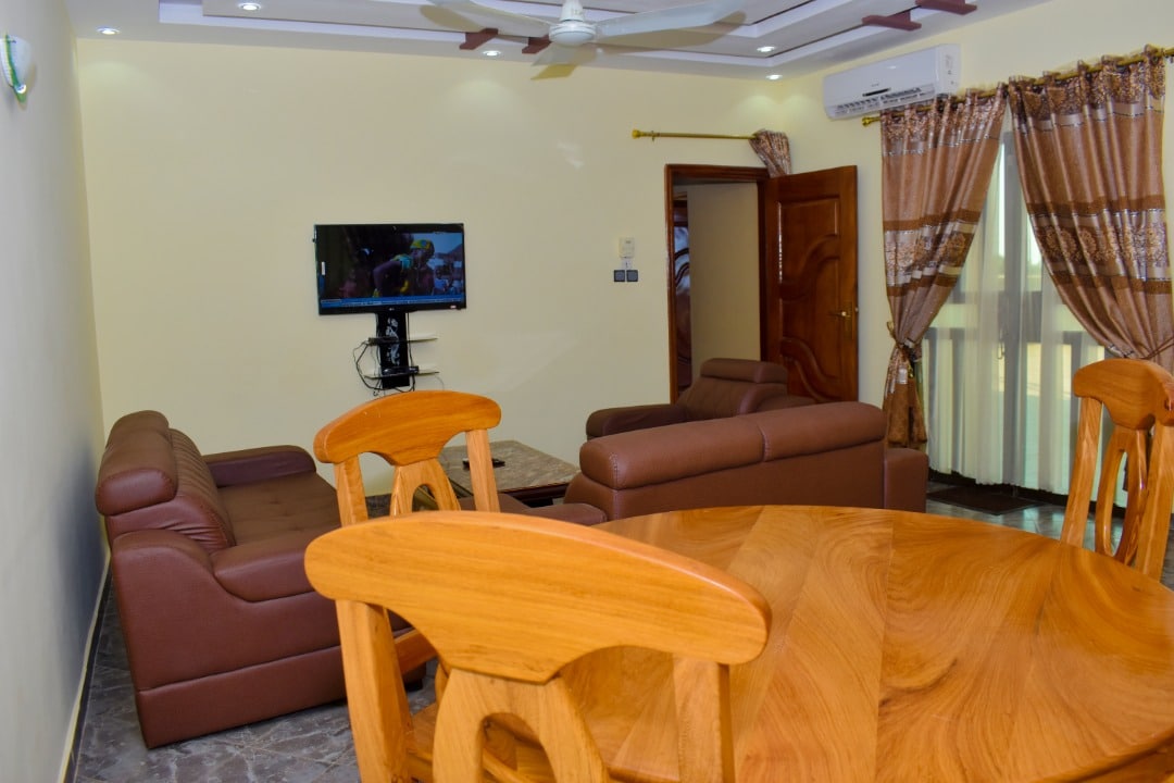 The spacious living and dining area features a wooden dining table surrounded by chairs. Two comfortable sofas are positioned for relaxation, facing a wall-mounted television. Natural light enters through large windows adorned with elegant curtains.