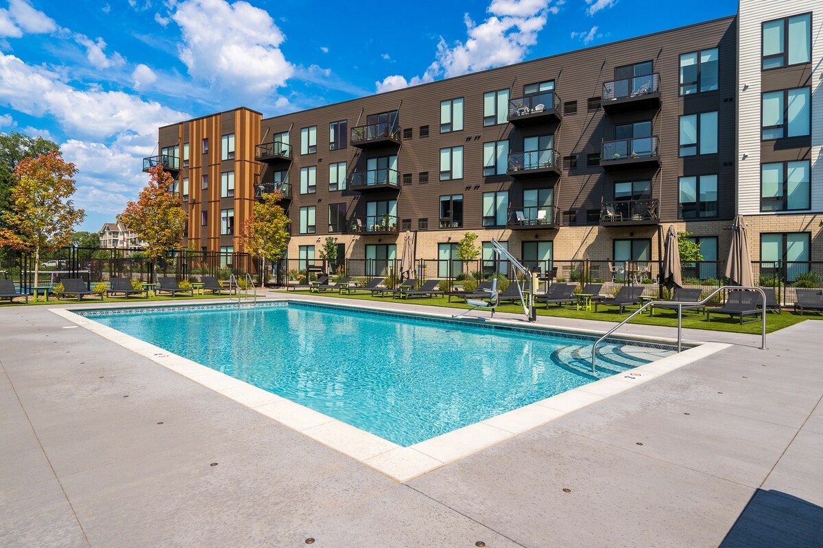 A community pool is surrounded by landscaped areas with lounge chairs. The modern building features multiple balconies, with large windows allowing for ample natural light. The sky is clear, highlighting a vibrant atmosphere in the outdoor space.