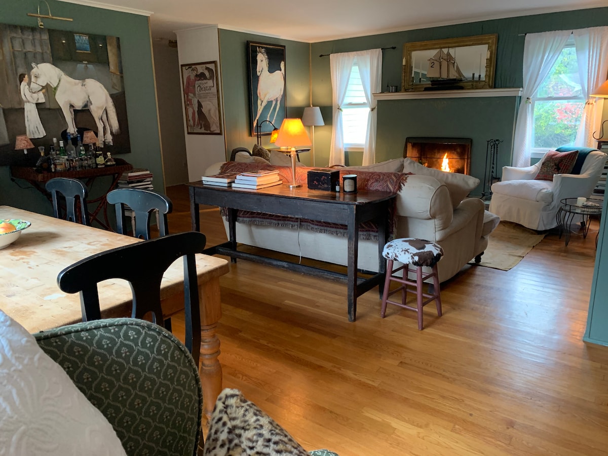 A warm and inviting living space features a comfortable sofa facing a wood fireplace, with a cozy armchair nearby. Decorated walls showcase equine artwork, and a side table supports a lamp. Natural light filters through sheer curtains over the windows.