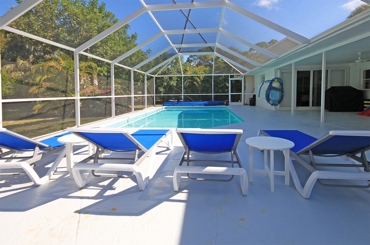 The expansive outdoor area features a screened lanai with a large heated pool. Several lounge chairs with blue cushions surround the pool, accompanied by a small white side table. Lush greenery is visible in the background, creating a tranquil setting.