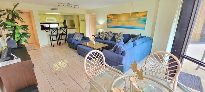 Relax in this bright open‑concept living area featuring a plush sectional, modern entertainment setup, and a sleek kitchen with bar seating. Perfect for gathering, unwinding, or cooking together, it offers comfort and style in your coastal retreat.