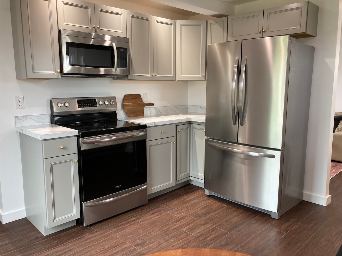 The kitchen showcases modern stainless steel appliances, including a refrigerator and stove. Light grey cabinets line the walls, complemented by marble countertops, creating a sleek and functional cooking space. The wood-look flooring adds warmth to this inviting kitchen environment.
