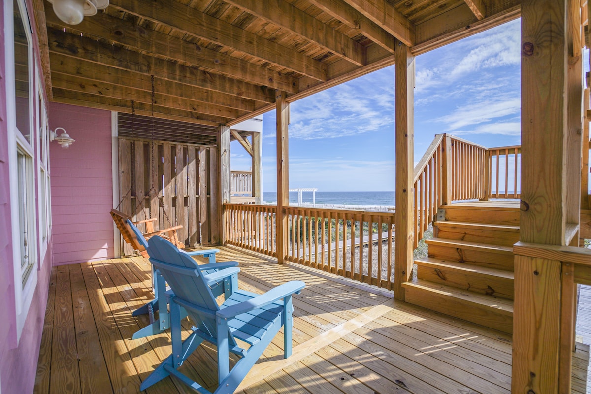 A spacious deck features wooden flooring and comfortable adirondack chairs, offering unobstructed views of the ocean. Wooden stairs lead up to the deck, enhancing accessibility to the outdoor space. Soft clouds are visible against a bright blue sky, creating a serene outdoor atmosphere.
