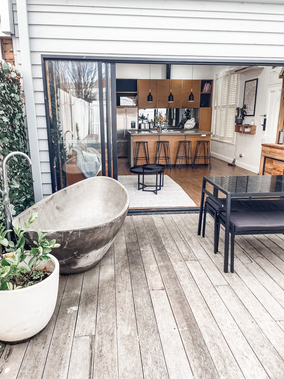 The outdoor area features a spacious deck with a deep concrete bath surrounded by greenery, a dining table set for four, and a view of the open-plan kitchen through large glass doors. Natural light highlights the warm wooden tones inside.