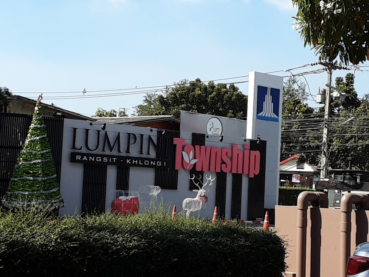 The entrance to the Lumpini Township is displayed, featuring a sign with the name 'Lumpin Rangsit Khlong 1' and 'Township' in contrasting colors. Nearby, a green Christmas tree ornament and a few holiday decorations are positioned alongside the entrance, with a clear blue sky overhead.