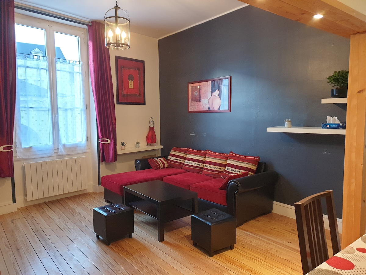 A cozy living area features a red couch with decorative cushions and a black coffee table. Large windows provide natural light and are framed by deep red curtains. A dark accent wall adds depth, while wooden flooring contributes to the warm ambiance.