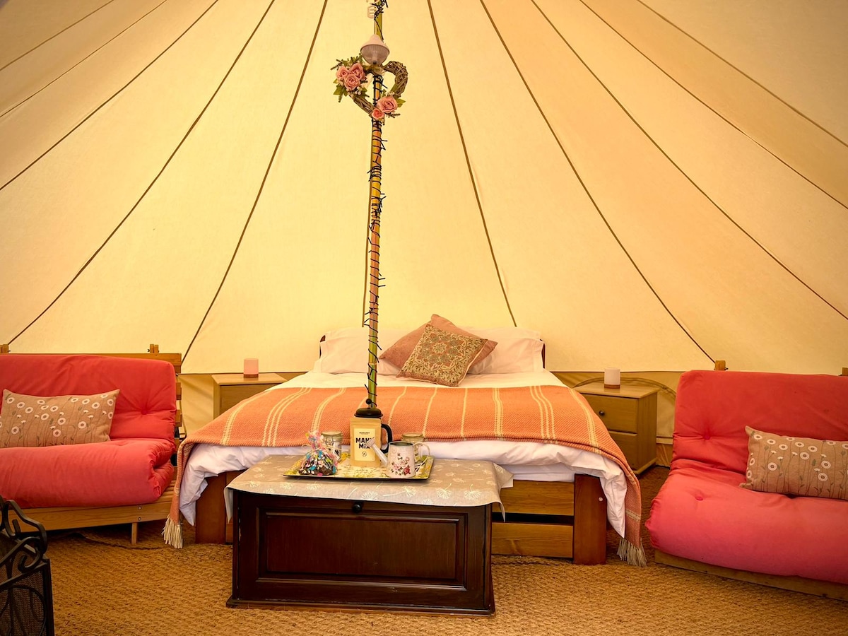 An inviting bell tent interior features a spacious bed adorned with a light blanket and decorative pillows. Two cozy futons flanking a wooden coffee table create a comfortable lounging area. Soft lighting and a floral accent piece at the tent's center enhance the tranquil ambiance.