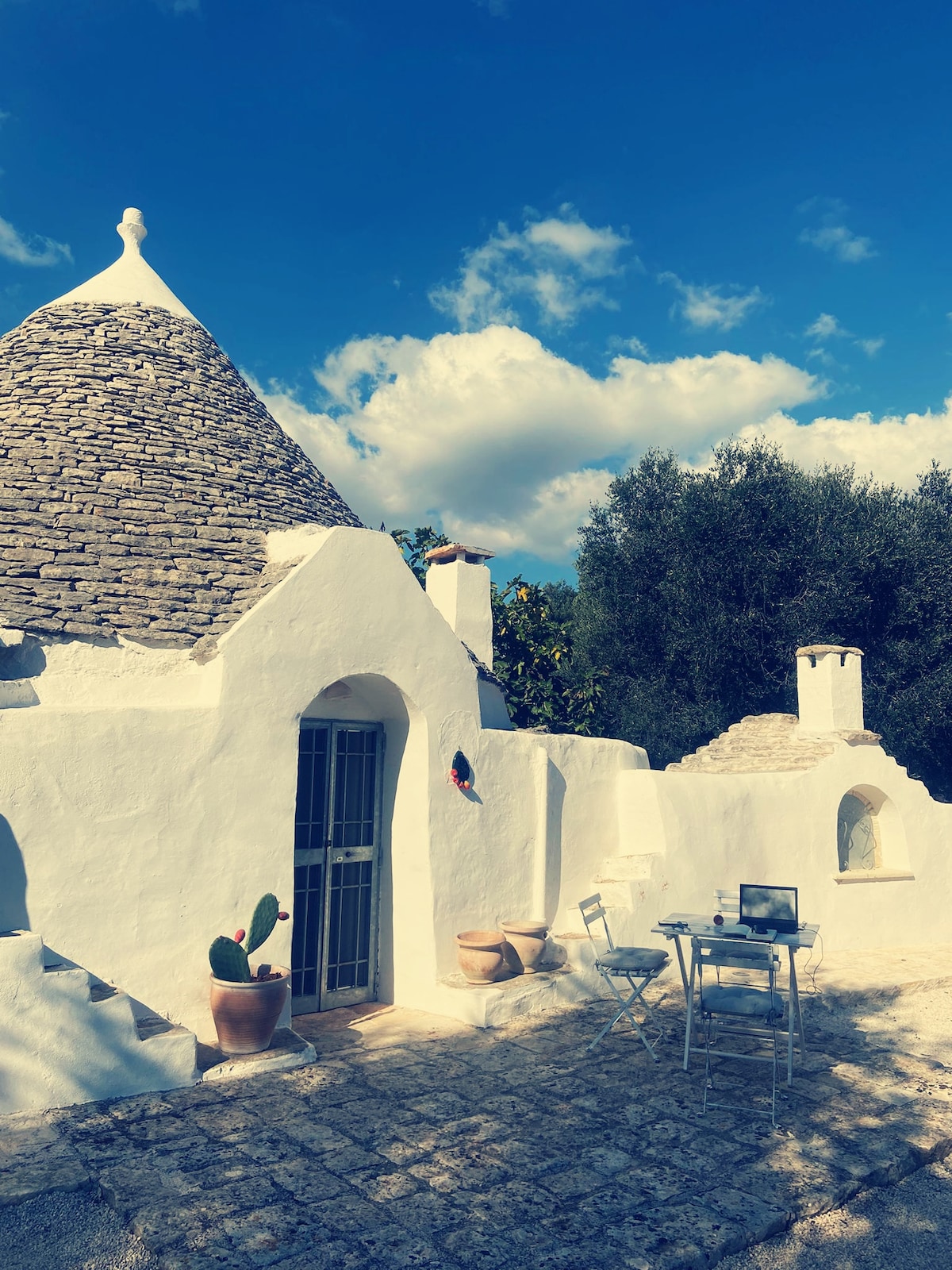 An exterior view of a traditional trullo showcases its iconic conical roof and whitewashed walls. A small table and chairs are positioned outside, surrounded by potted plants. The clear blue sky and fluffy clouds create a serene backdrop.
