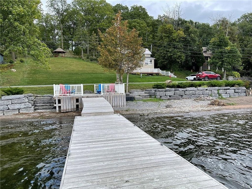 A wooden dock extends into the calm waters, bordered by a grassy area that features a small gazebo and a cottage in the background. Stone retaining walls frame the shoreline, while colorful chairs are positioned at the dock, offering a place to relax and enjoy the scenery.