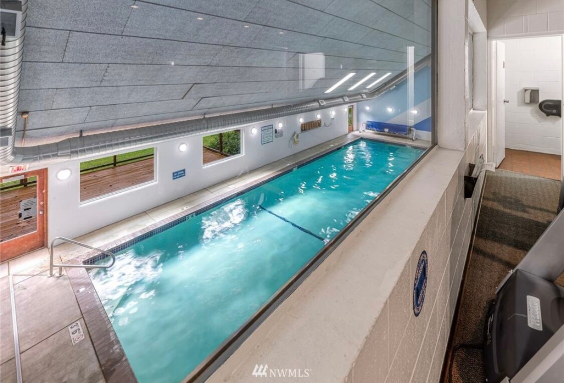 An indoor heated swimming pool is visible through large windows, with water gently rippling under soft lighting. The pool area is well-maintained, featuring tile walls and a seating area nearby. Access to restrooms is also available.