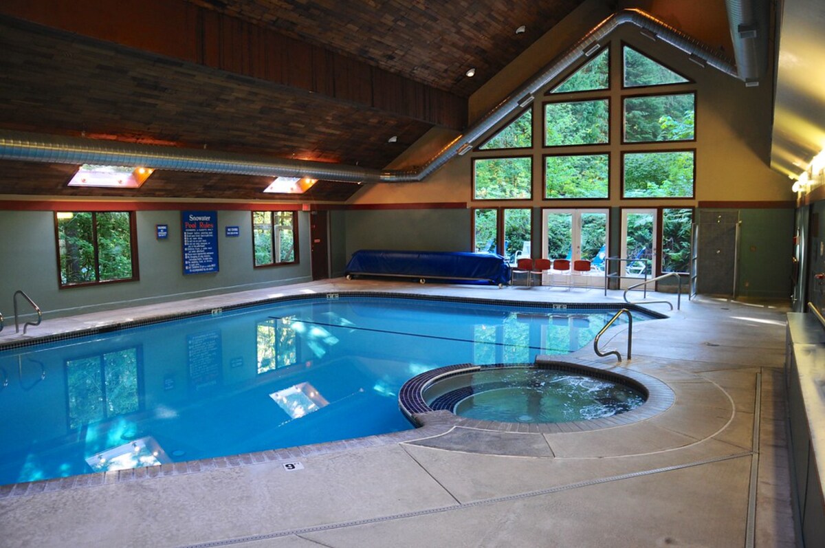 An indoor pool area is seen featuring a large swimming pool complemented by a separate hot tub. High ceilings with wooden beams create an airy atmosphere. Large windows bring in natural light and showcase views of the surrounding greenery. Lounge chairs are placed nearby.