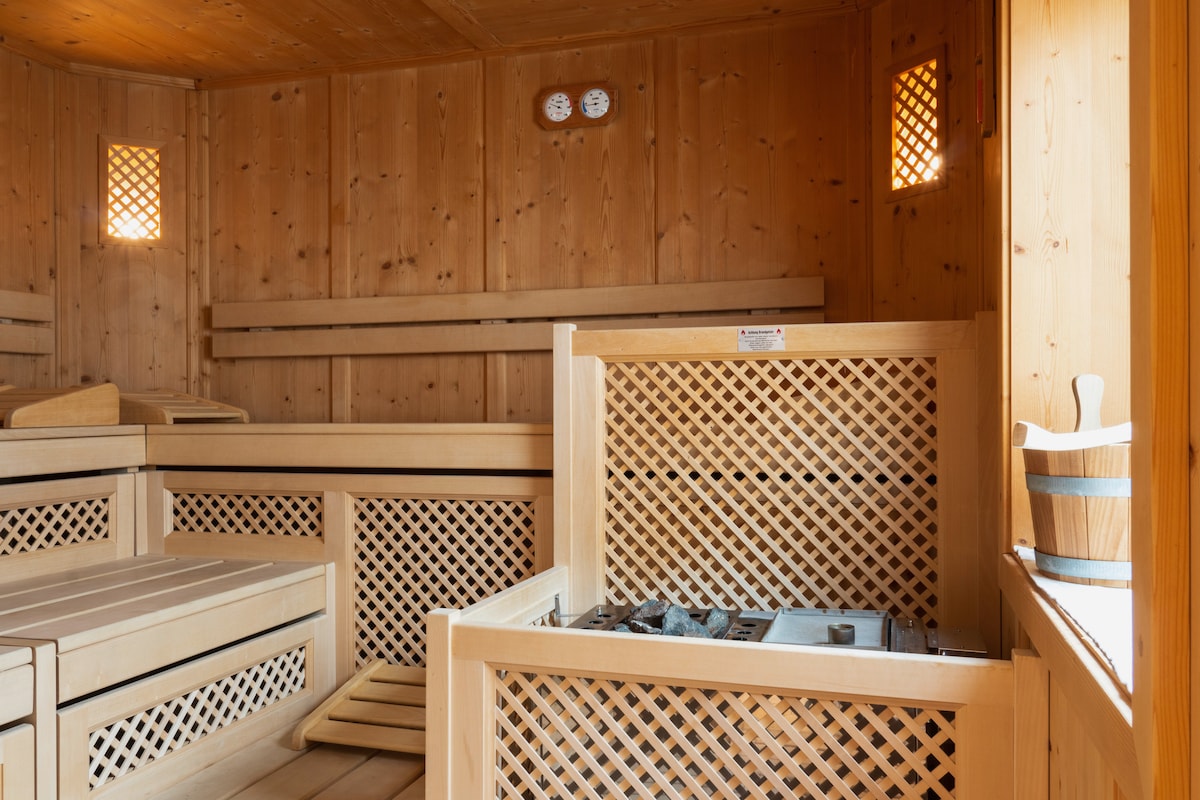 A wooden sauna interior is observed, featuring a series of benches and a stove with a stone grate. Small windows provide soft light, and wooden lattice walls create a warm ambiance. Thermometers are mounted on the wall, indicating temperature.