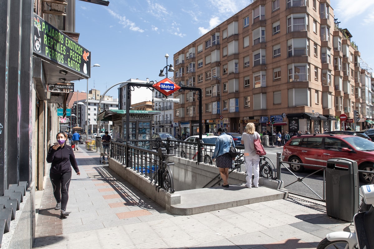 A bustling city street is depicted, showcasing the entrance to a metro station marked by a prominent blue 'Metro' sign. Surrounding buildings display a mix of residential and commercial spaces, while pedestrians navigate the sidewalk on a sunny day.