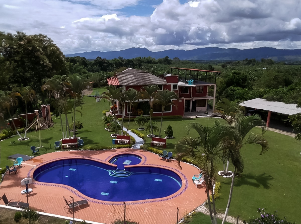 A spacious outdoor area features a large heart-shaped swimming pool surrounded by lounge chairs and palm trees. A red two-story building is visible in the background, with mountain views and green grass extending throughout the landscape.