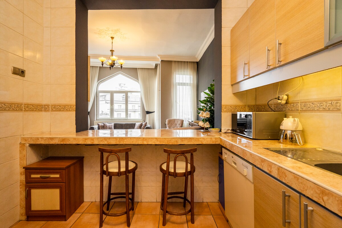 A well-equipped kitchen is showcased with modern appliances and a spacious countertop. Two wooden stools are positioned near the counter. Natural light illuminates the area, highlighting the cream-colored tiles and cabinetry, while a view into the living room is visible through the archway.