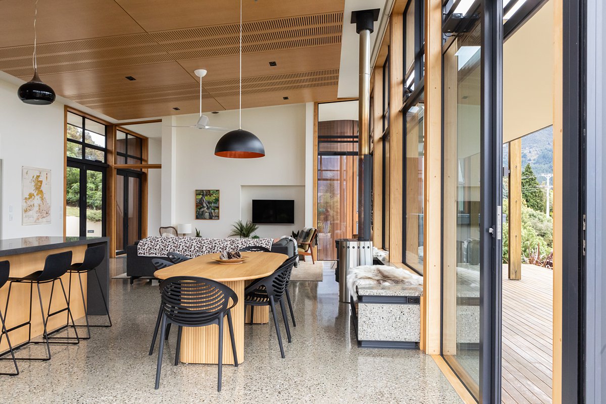 An open plan living area showcases large glass doors that connect the indoor space with an outdoor deck. Modern furniture includes a spacious dining table with black chairs and a cozy seating arrangement, complemented by polished wooden panels and a warm color palette.