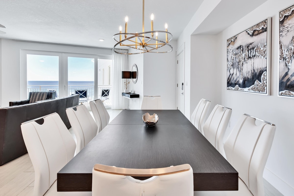 A spacious dining area features a large dark table surrounded by eight white chairs. Large glass doors provide an unobstructed view of the ocean, and a modern chandelier hangs overhead. Two artworks adorn the adjacent wall, complementing the bright and airy design.