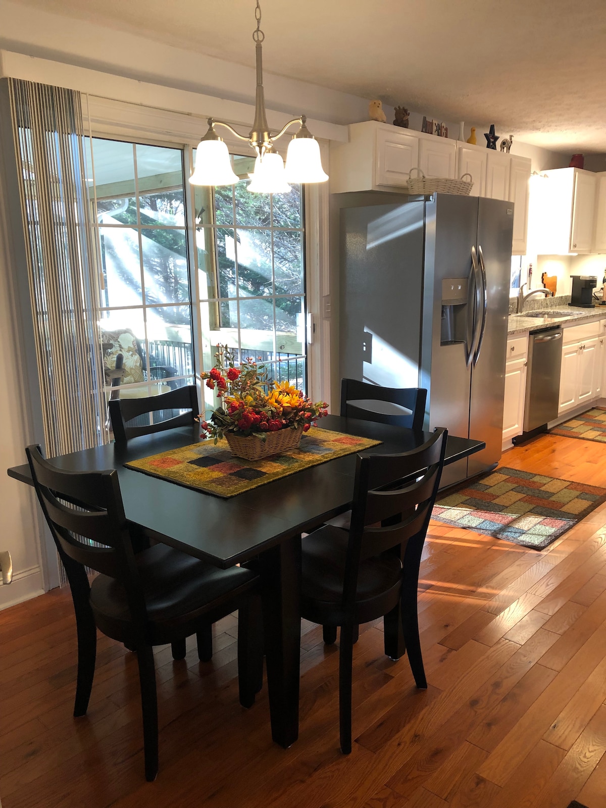 A dining area is presented with a small black table and four matching chairs. A seasonal centerpiece is placed on the table, surrounded by natural light from large windows. A modern refrigerator is visible in the background, alongside bright cabinetry and hardwood flooring.