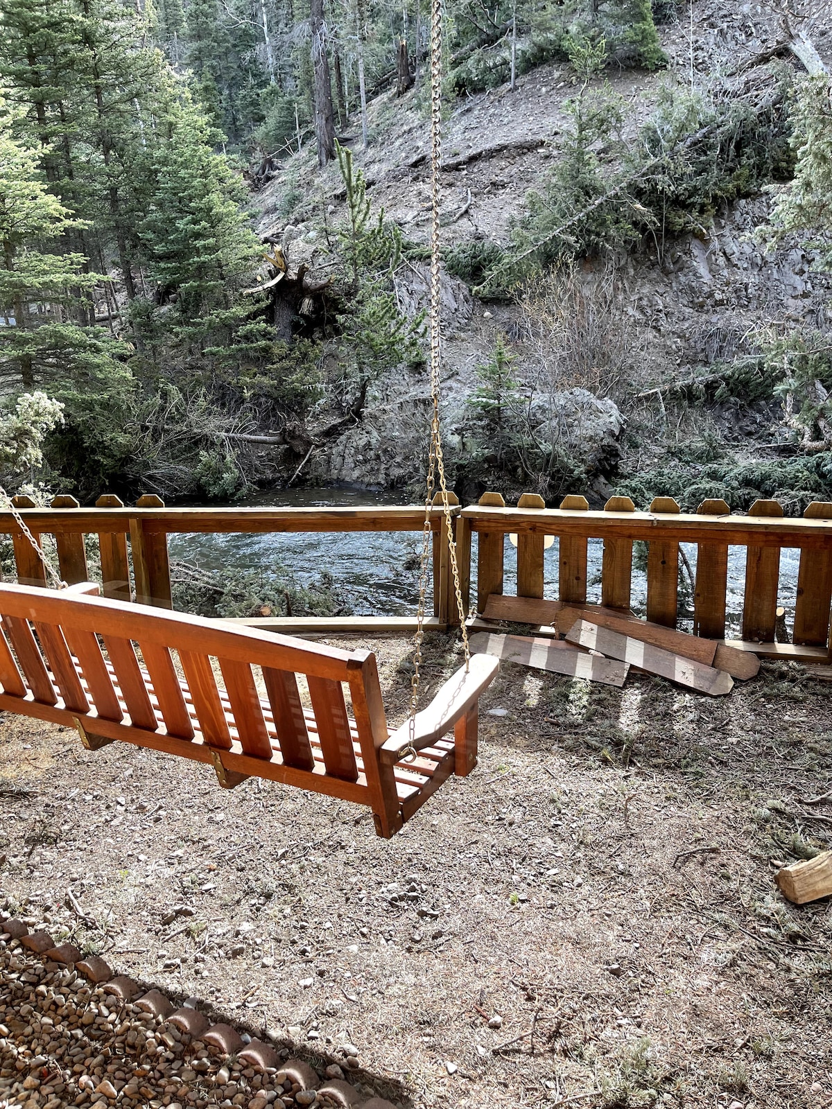 A wooden bench swing is situated on a patch of natural ground beside a gently flowing river. Surrounding trees provide shade, creating a serene outdoor space for relaxation. The swing is positioned against a rustic wooden fence that complements the natural setting.