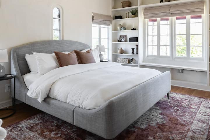 Ზემოთ საძინებელი, რომელშიც არის საწოლი „King Saatva“, 55"-იანი ტელევიზორი, 640 ‑ იანი თეთრეული, კარგად მოპირკეთებული წიგნების თაროები და უშუქო ფანჯრის ფერები.