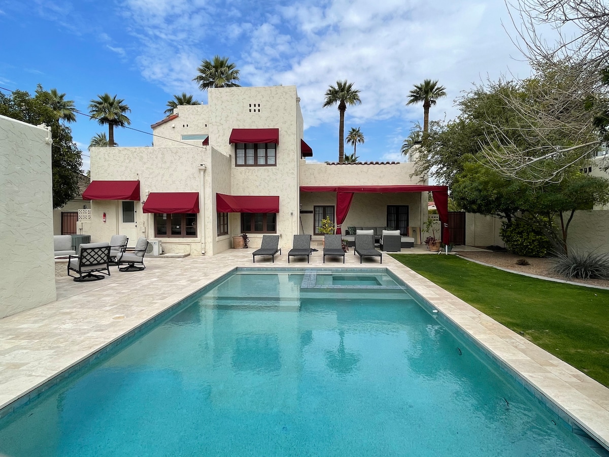 The outdoor area features a crystal-clear pool surrounded by stone tiles, with comfortable lounge chairs positioned on one side. A shaded seating area is visible under a fabric awning, providing a relaxing atmosphere. Palm trees line the backdrop, enhancing the serene setting.