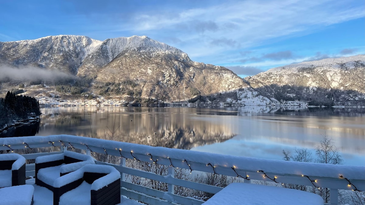 The outdoor terrace offers a panoramic view of snow-covered mountains reflecting in the calm fjord waters. Cozy seating arrangements are visible, with soft lighting enhancing the serene atmosphere. The clear blue sky contrasts with the snowy landscape, creating a tranquil winter scene.