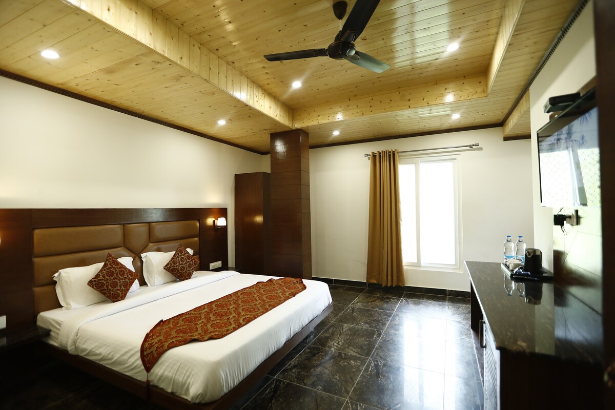 A well-appointed room is equipped with a spacious bed adorned with decorative pillows. A sleek wooden ceiling complements the room's modern design. Natural light enters through a window dressed with soft curtains, while a flat-screen television and a dark console table add functionality to the space.