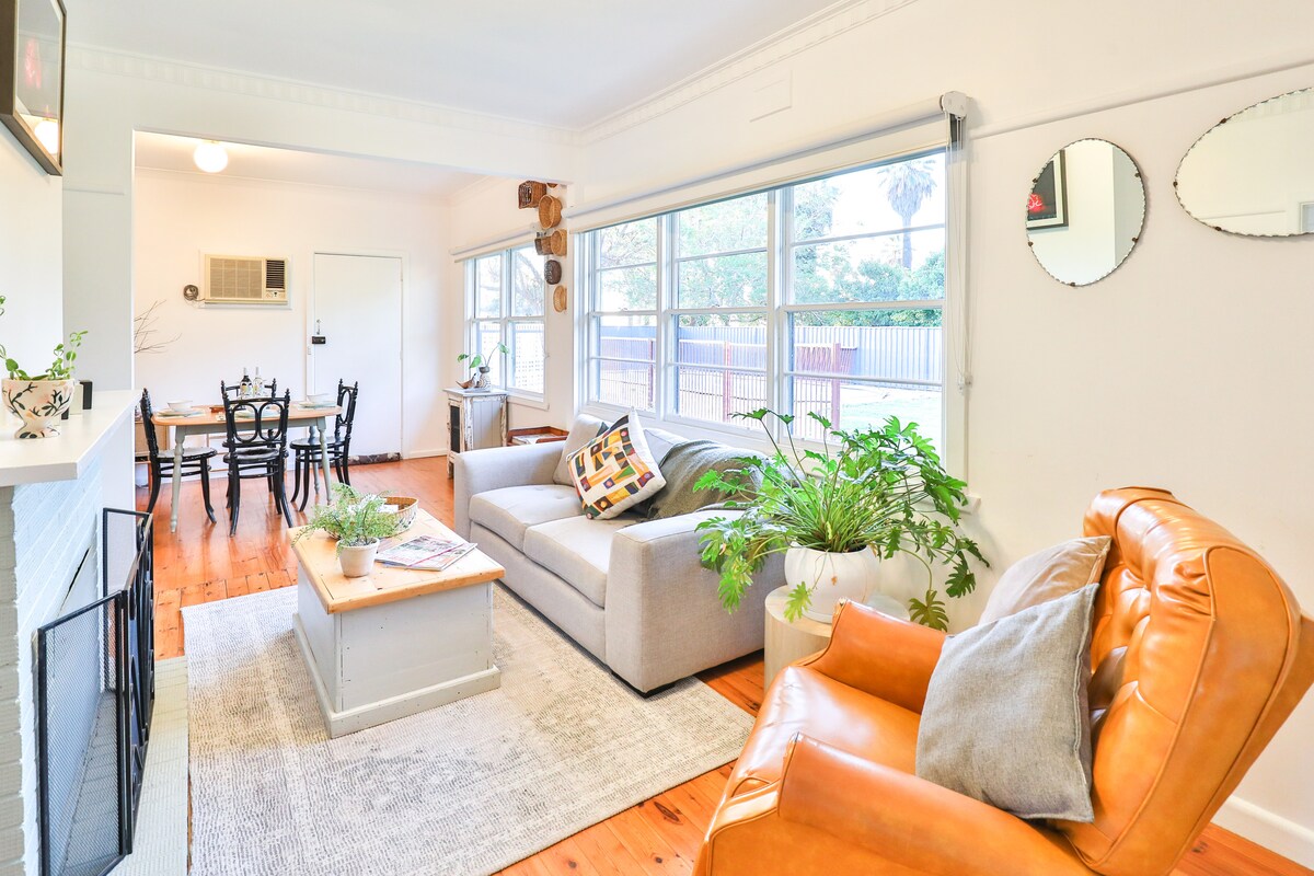 A cozy living area is highlighted by a comfortable gray sofa and a single brown leather chair. A light rug sits atop polished wooden floors, while a coffee table holds decorative plants. Large windows provide natural light and views of the outdoors.