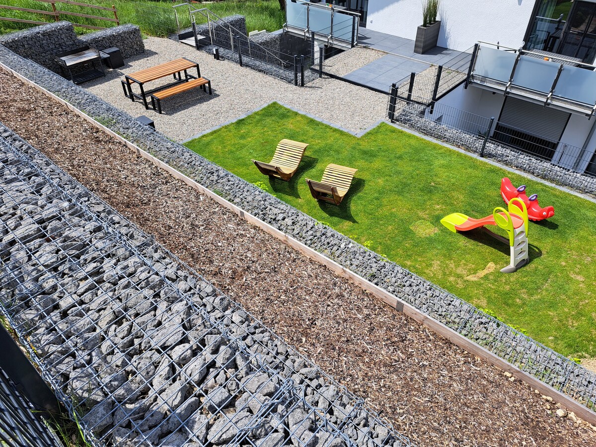 An outdoor area is presented with a well-maintained lawn and two lounge chairs facing each other. Tables are arranged on gravel, with a children's play structure featuring a slide and climbing elements prominently displayed. The space is surrounded by a natural stone wall.