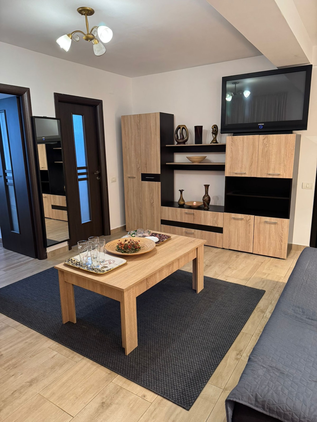 A modern living space features a wooden coffee table at the center, surrounded by a dark rug. Light-colored cabinetry and decorative items are displayed on shelves. A flat-screen television is mounted on the wall, and natural light is welcomed through a nearby door.