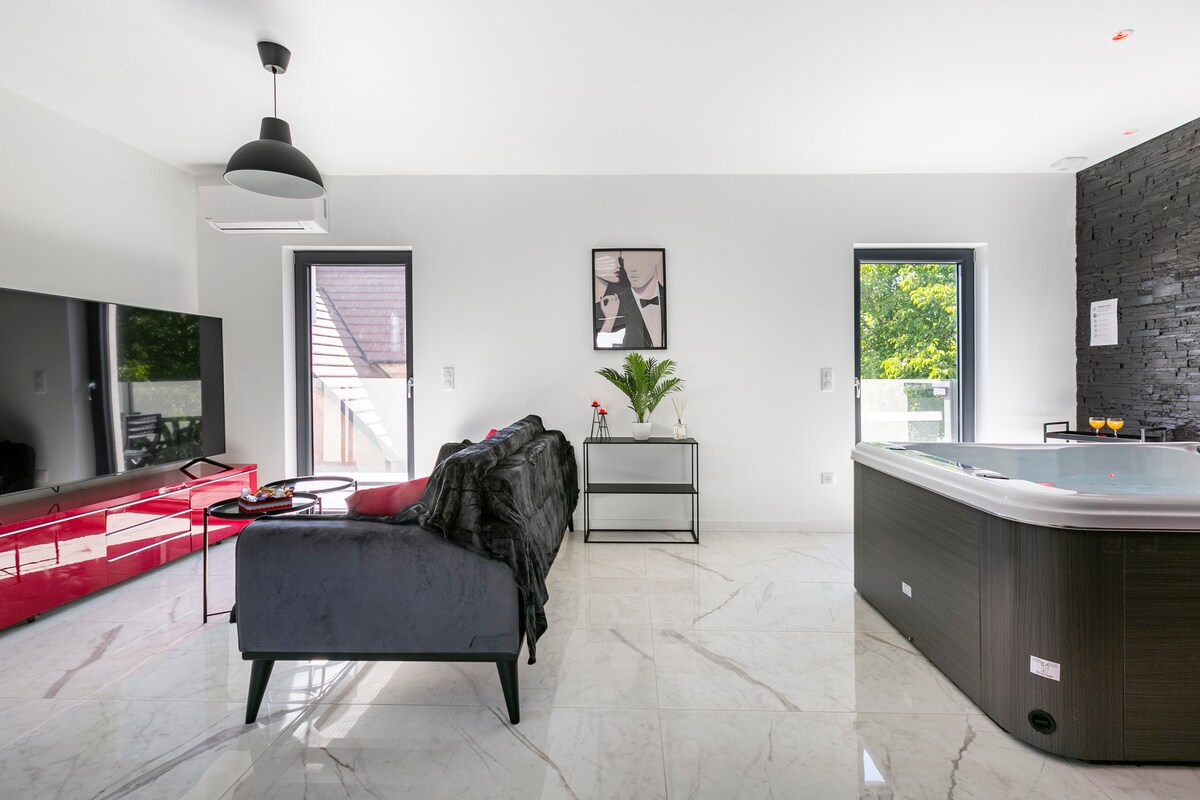 A modern living area features a spacious black sofa positioned near a large flat-screen TV. A private jacuzzi is visible to the right, accentuated by elegant tile flooring. Natural light enters through windows, highlighting a minimalistic artwork and decorative plant on a side table.