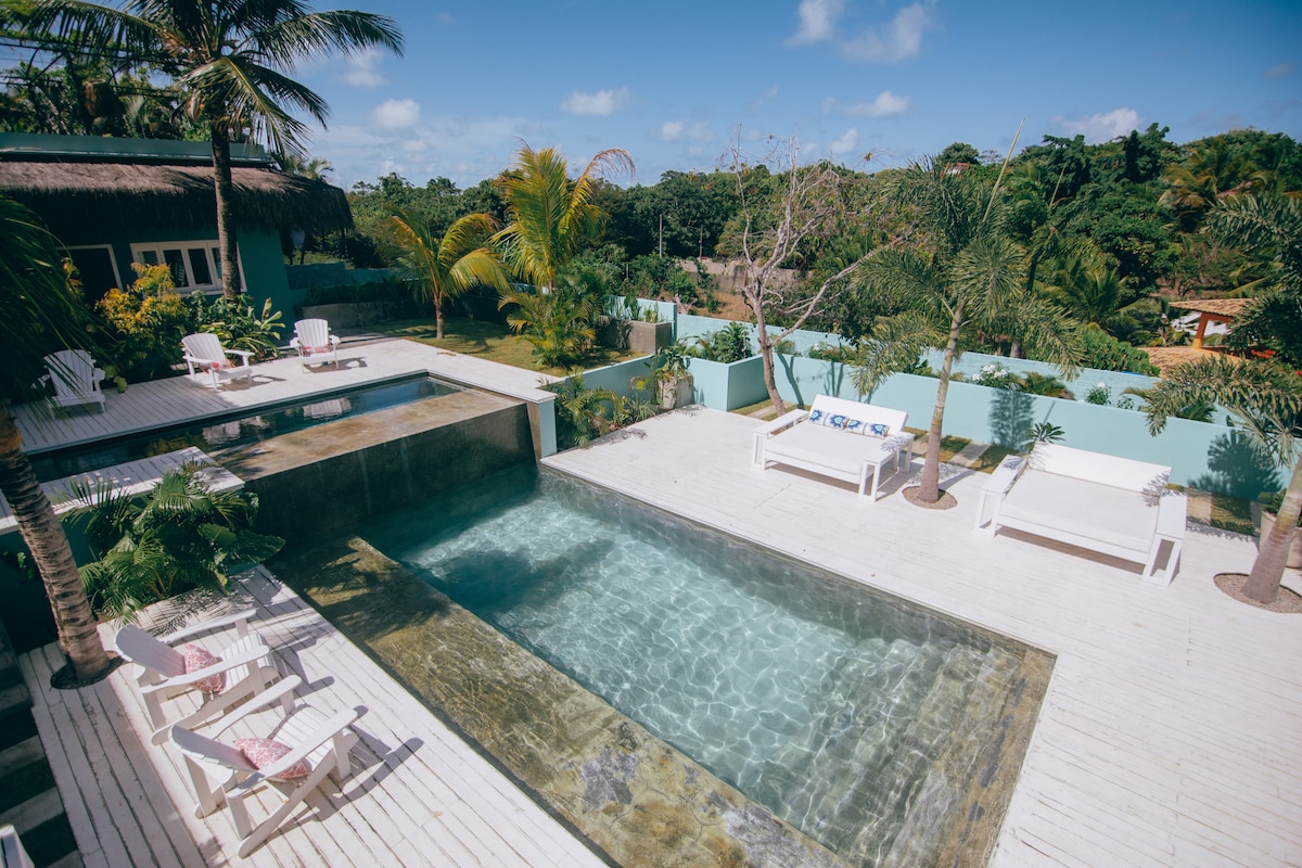 An outdoor area is showcased featuring a shared pool surrounded by a wooden deck. Sun loungers and seating areas are positioned under palm trees, with lush greenery in the background. The clear water of the pool reflects the bright blue sky, creating a serene atmosphere.