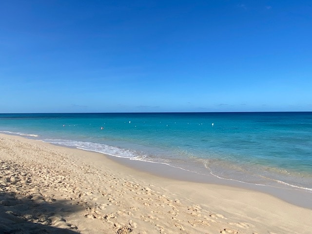 A sandy beach stretches along crystal clear waters, transitioning from light turquoise to deep blue. Gentle waves roll onto the shore, leaving delicate ripples in the sand. Bright skies enhance the serene atmosphere, with a few buoys visible offshore.
