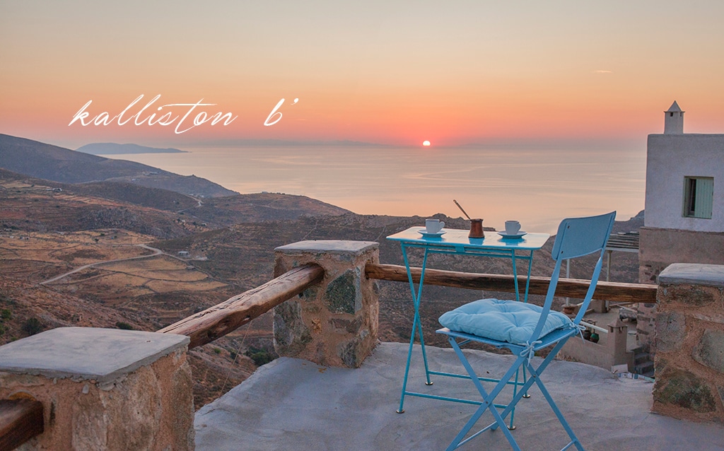 A small outdoor seating area features a light blue table and chairs, set against a backdrop of a vibrant sunrise over the Aegean Sea. The landscape displays gentle hills and valleys, highlighting the serene natural beauty of the surrounding area.