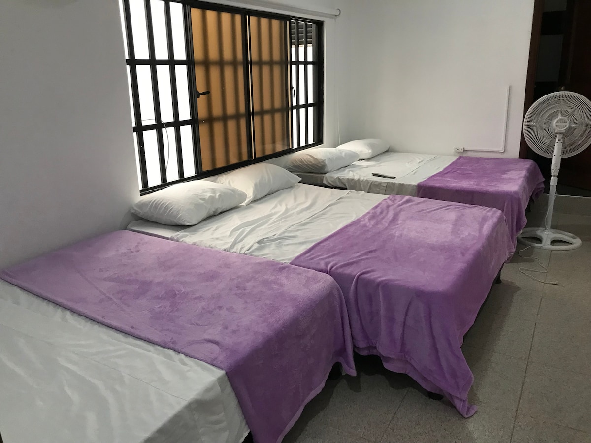 The sleeping area features three beds, each covered with purple throws. Spacious pillows are arranged on the beds. A window with vertical black bars allows natural light to enter, while a standing fan is positioned nearby for ventilation.