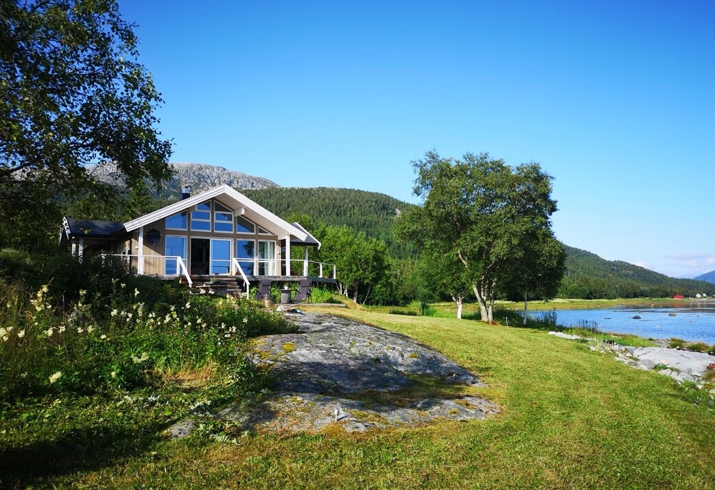 A modern cabin is situated near a tranquil body of water, surrounded by green grass and rocky terrain. Large windows reflect natural light, offering views of the landscape. Lush trees and distant mountains create a serene backdrop for this peaceful getaway.