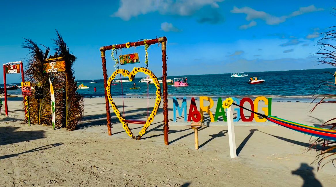 A sandy beach scene is displayed with colorful signs creating a lively atmosphere. A heart-shaped decorative frame is prominently featured, surrounded by vibrant plants. Several small boats can be seen on the calm waters, contributing to the relaxing coastal environment.