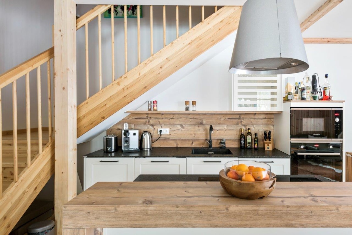 The kitchen features natural wood accents and modern appliances, including a microwave and a coffee machine. A stone countertop holds a bowl of fresh fruit, while the open space is illuminated by a contemporary lampshade, enhancing the inviting atmosphere.