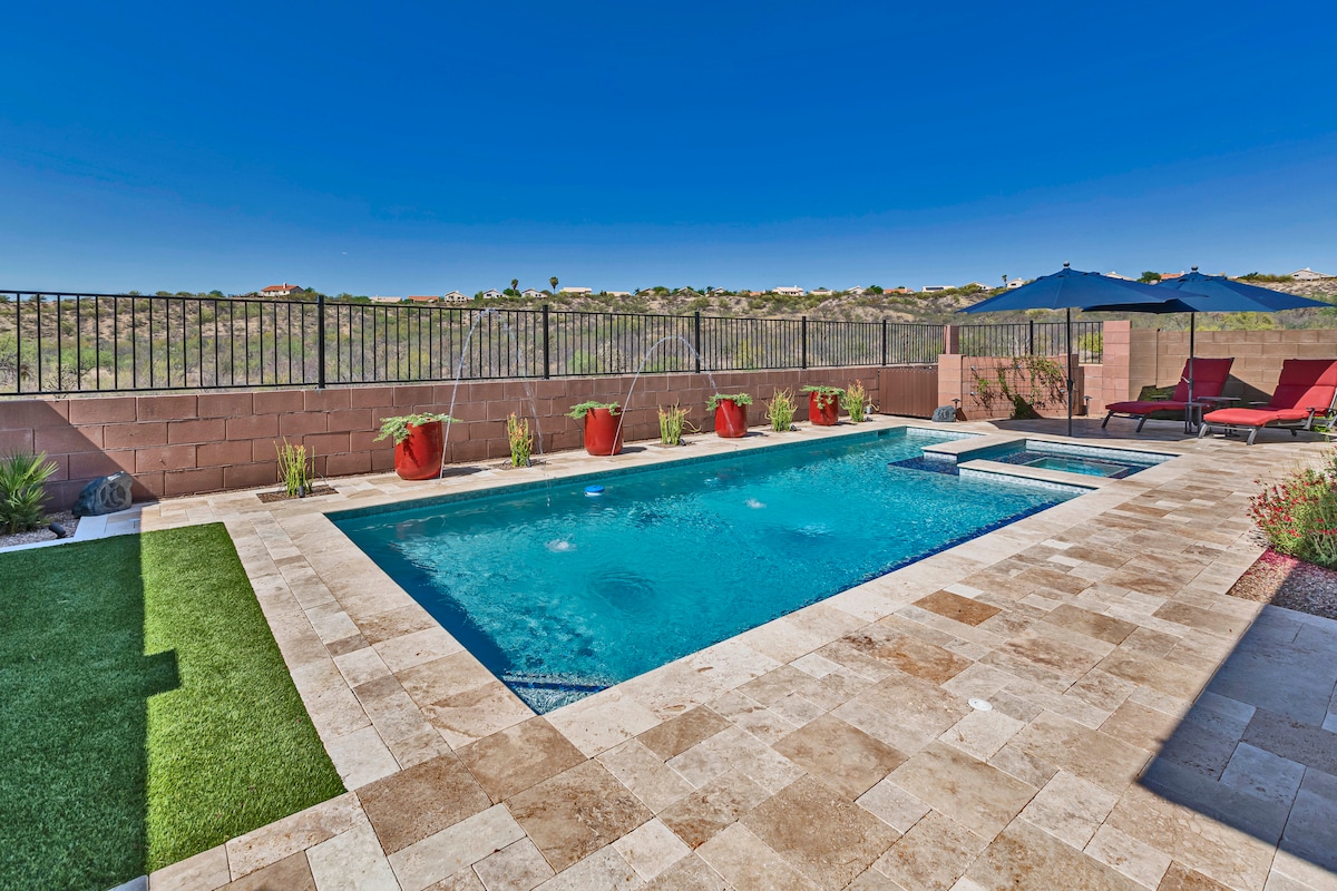 A private pool is surrounded by travertine hardscape, featuring vibrant red pots with flowering plants. Lounge chairs are positioned near the water under shaded umbrellas, with a low fence providing an unobstructed view of the landscape beyond.