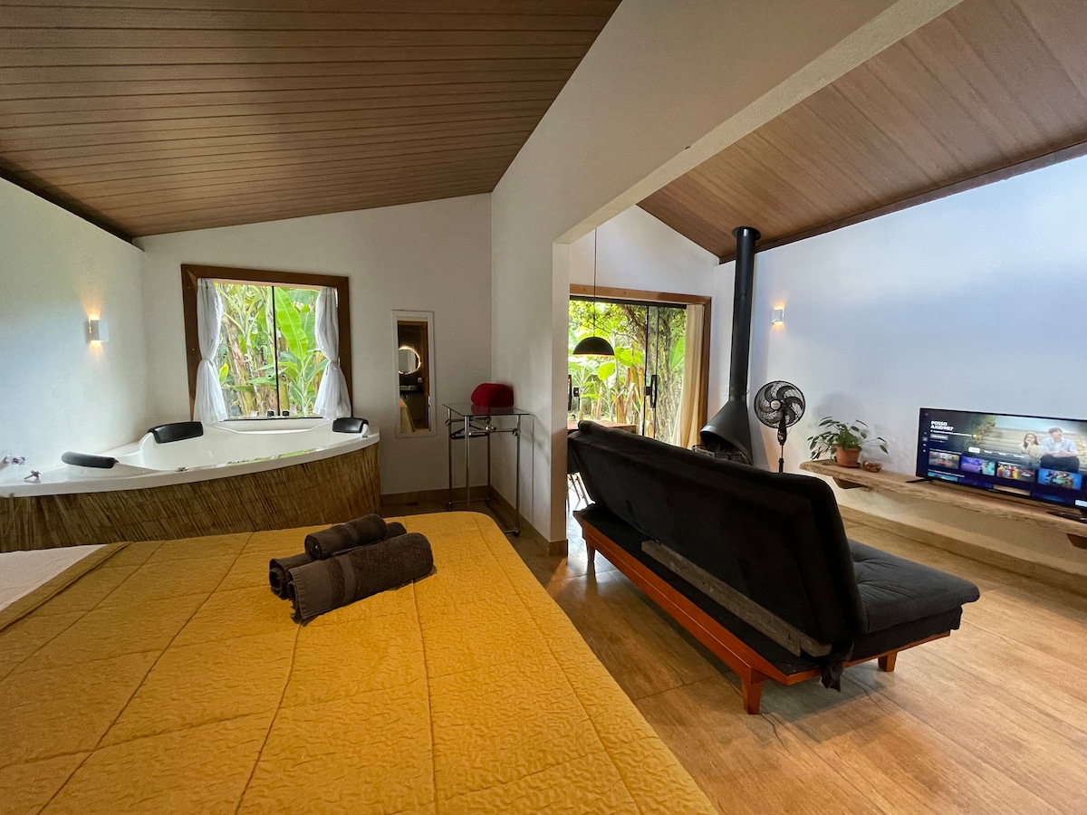 A cozy interior space is showcased, featuring a large bed covered with a golden blanket. A black sofa faces a mounted television, while a mini spa is located beside large windows that provide views of lush greenery outside.