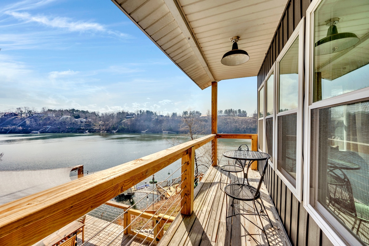 A wooden deck extends over the water, featuring two black wire chairs and a small table. Panoramic views of the lake and surrounding landscape are visible, with the shoreline and distant trees reflecting the tranquil setting.