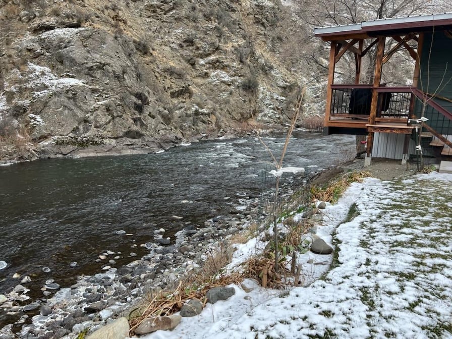 The Little Salmon River flows gently beside the cabin, framed by rocky terrain and sparse vegetation. A wooden deck extends from the cabin, offering views of the water and surrounding landscape. Snow lightly covers the ground, contrasting with the river's surface.