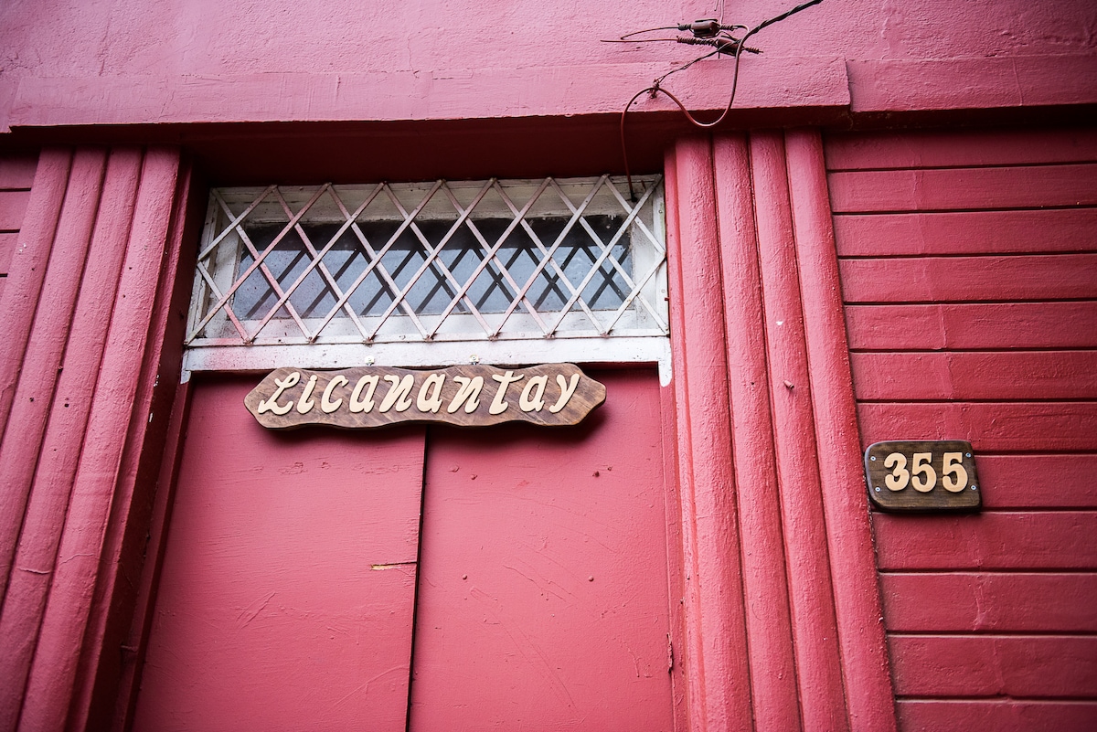A red exterior wall features a wooden sign reading 'Licantantay,' positioned above a pronounced door. A decorative grate covers a small window above the entrance. The door is framed by vertical panels, with the number '355' displayed prominently on the right side.