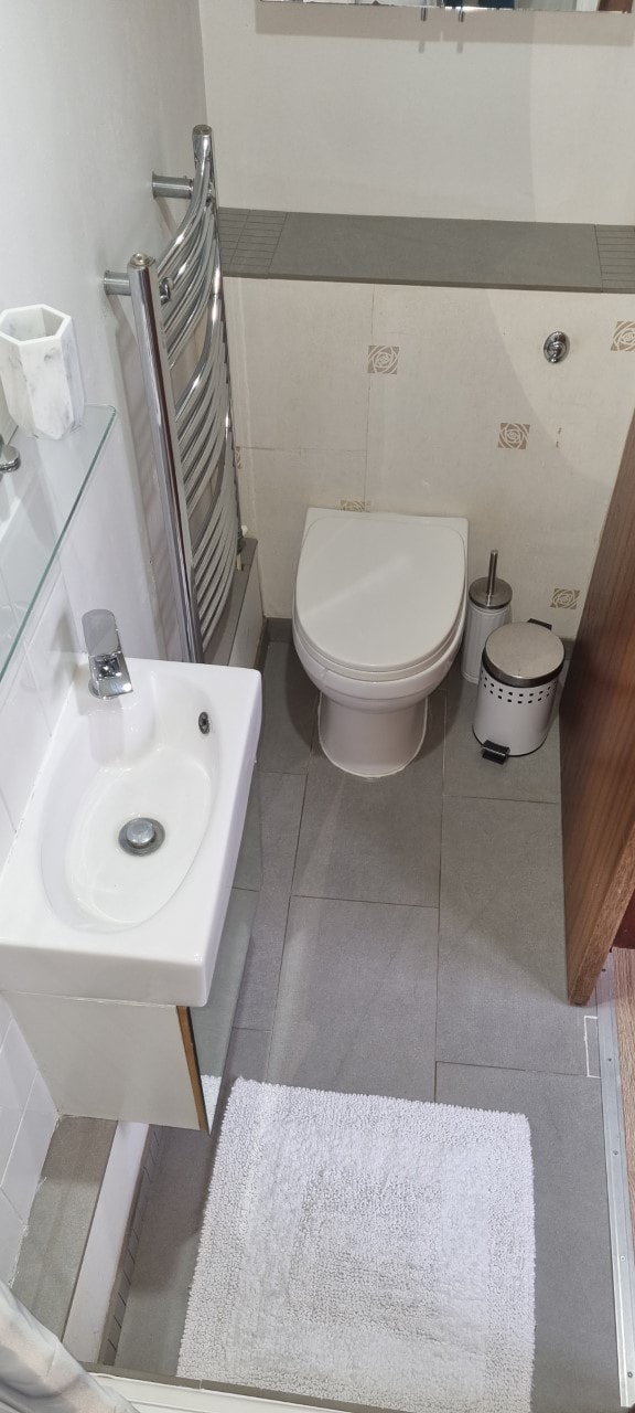 A compact private bathroom is depicted, featuring a modern toilet and a small sink with a chrome faucet. A wall-mounted towel radiator adds functionality. Soft gray tiles cover the floor, complemented by a white bath mat. A mirror is visible above the sink.